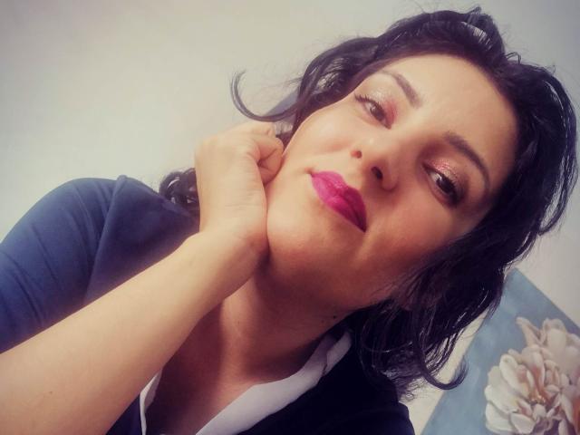 AddictedToUTS with dark hair, pink lipstick, and shiny eyeshadow, resting her chin on her hand, wearing a blue shirt and white collar, looking at the camera.