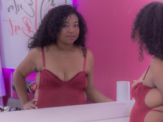 XairaRuiz with curly black hair, wearing a red spaghetti-strap dress, standing in front of a mirror, looking at herself, showing her tits, in a pink-lit room.