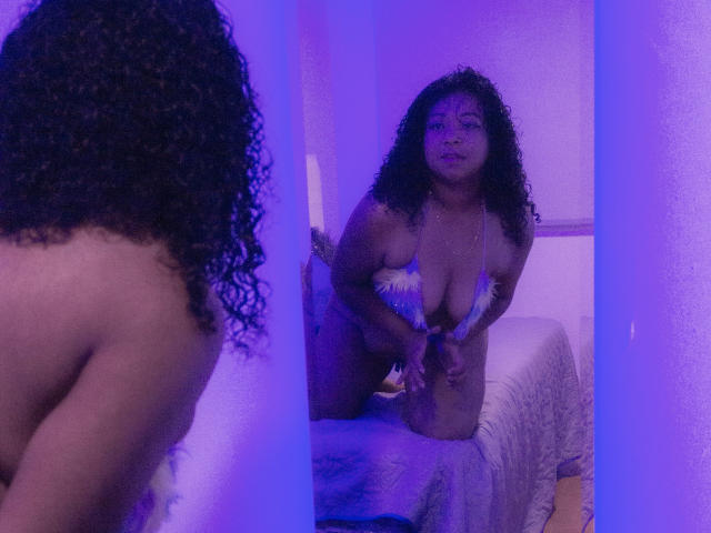 XairaRuiz, con i capelli neri ricci, la pelle marrone media, il seno abbondante e un bikini floreale, è inginocchiata di fronte a uno specchio con illuminazione viola, che riflette la sua schiena nuda..