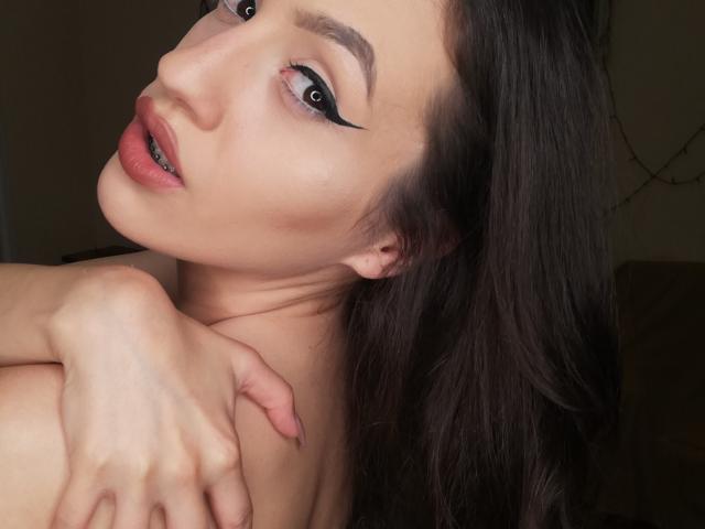 Vievienne Clemente, avec ses cheveux foncés, son eye-liner noir, ses lèvres roses et son sourire séduisant, tient son épaule nue, dévoilant sa poitrine, avec un regard doux, séduisant et confiant..