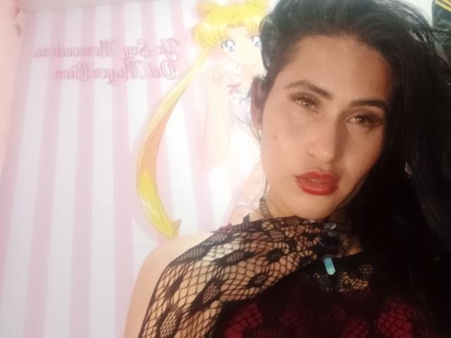 ElectraHottyX with black hair, red lipstick, and a fishnet top, showing her tits slightly, next to a pink-striped wall and a Sailor Moon poster.