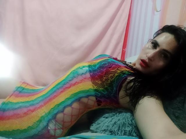 ElectraHottyX with wet black hair, red lipstick, wearing a rainbow-striped fishnet bodysuit, lying on a green pillow, exposing her tits through the net. Her face is slightly blurred.