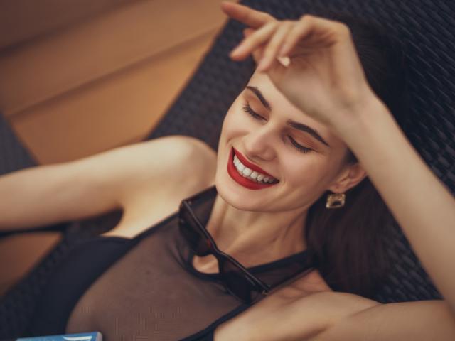 BatSama with closed eyes, red lipstick, black sheer top, and sunglasses on chest, smiling, hand covering forehead, showing her tits slightly, lying on dark wicker chair.