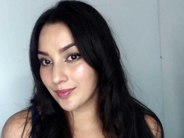 Julianal with black hair, brown eyes, and pink lips, wearing a dark sleeveless top, has a slight smile and a smooth, fair-skinned face.