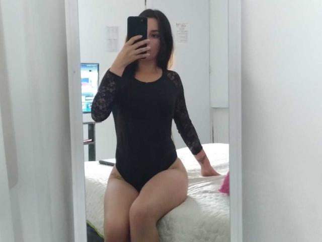 Julianal with dark lace long-sleeve black bodysuit, sitting on white bed, holding phone, showing thick thighs, small butt, and medium-sized tits, in mirror selfie.