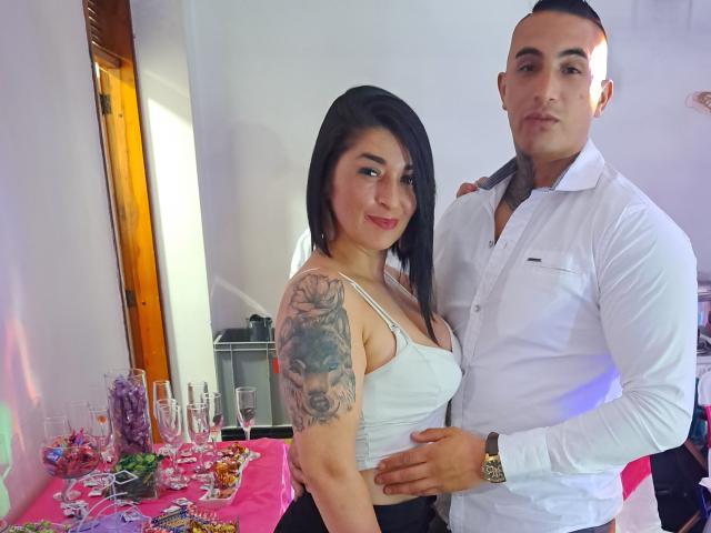 KarenAndScottI, com cabelo escuro, camiseta branca e tatuagem no braço direito, está ao lado de ScottI, que veste uma camisa branca e segura a cintura dela. No fundo, há uma mesa com copos e doces..
