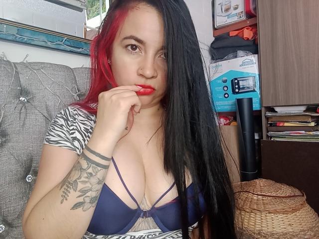 PAMDORA met zwart en rood haar, rode lippen, een top met zebravlekken, die haar borsten laat zien in een blauwe bh, een getatoeëerde arm, een vinger dichtbij haar mond, op een grijze bank, met een rieten mand..