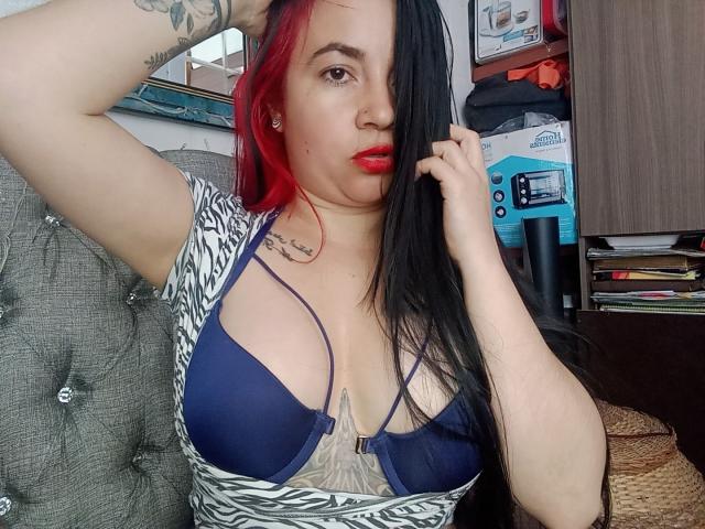 PAMDORA, avec des cheveux roux et noirs, portant un soutien-gorge bleu, montrant de grandes seins, une chemise à motifs zébrés, un tatouage sur le bras droit, une main dans les cheveux, du rouge à lèvres rouge, assise sur un canapé gris..