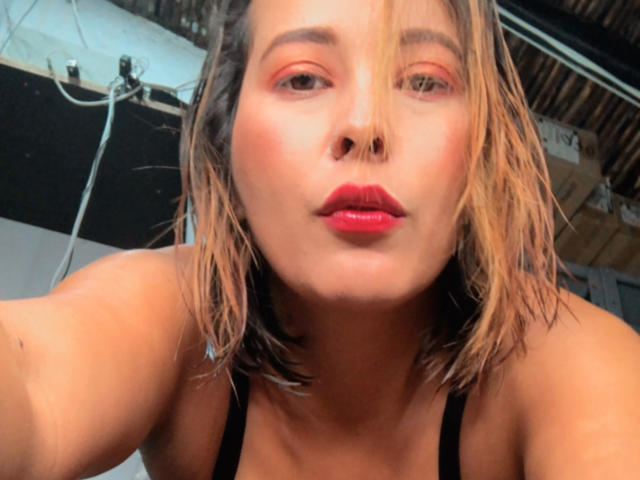SaritaVelez has dirty blonde, wet-looking hair, red lipstick, and subtle eye makeup. She's wearing a black strap top, showing her bare shoulders and a slightly open mouth.
