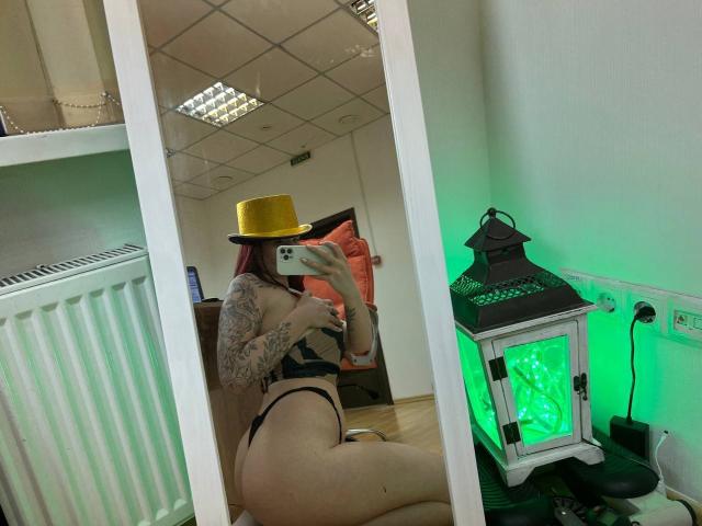 Innysik with a yellow hat, black lingerie, and tattooed arm, showing her bare ass in a mirror selfie, holding a phone, with a green-lit lantern on the right.