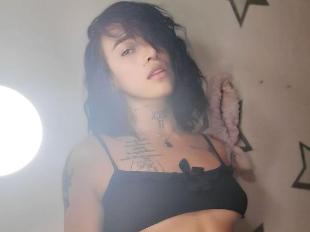 YeiSela with black hair, wearing a black bra, showing tattoos on neck and chest, looking at camera, light source on left, soft pink furry accessory on right.