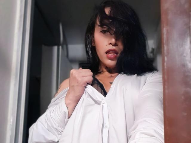 YeiSela with black hair, white shirt, and black bra, slightly open, pulling shirt down, showing a bit of her tits, with a teasing look, in a dimly lit room.