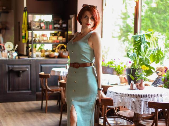 LisaTreds mit roten Haaren, die ein grünes Kleid mit Knöpfen und einen braunen Gürtel trägt, steht neben einem Tisch mit einer weißen Tischdecke aus Spitze und zeigt einen Ausschnitt in ihrem Kleid..