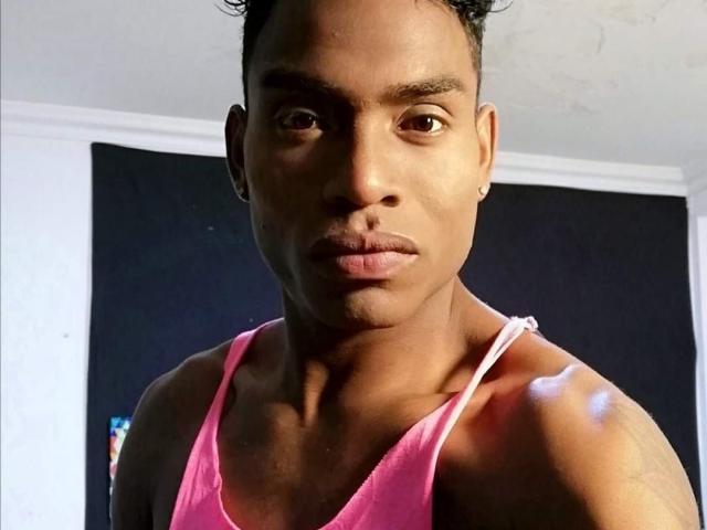 TheLordRox tiene un tono de piel marrón medio, cabello negro corto y rizado, ojos marrones, pequeños aretes, y lleva una camiseta rosa que deja al descubierto sus hombros y pecho..