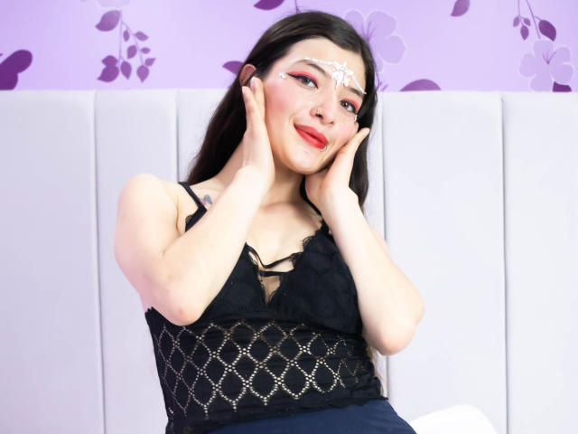 TeddyAndHottie with hands on cheeks, wearing black lace top, white facial paint, red lipstick, and dark hair, smiling, sitting on white cushioned surface.