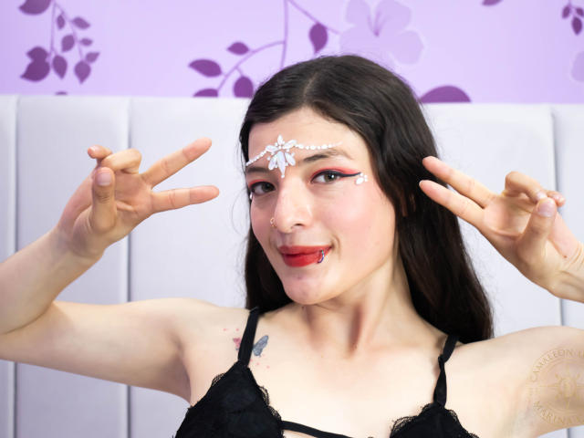 TeddyAndHottie con capelli scuri, indossa una maglia di pizzo nero, trucco sul viso con fiori bianchi, rossetto rosso, fa il segno della pace, ha un piccolo tatuaggio di una farfalla sulla spalla destra, pelle chiara, sorridente..