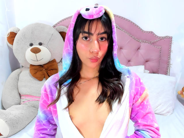 JessicaSoler with pink and purple tie-dye hood, wearing a unicorn costume, pouty face, visible cleavage, black hair, and pink cheeks, standing next to a large gray teddy bear.