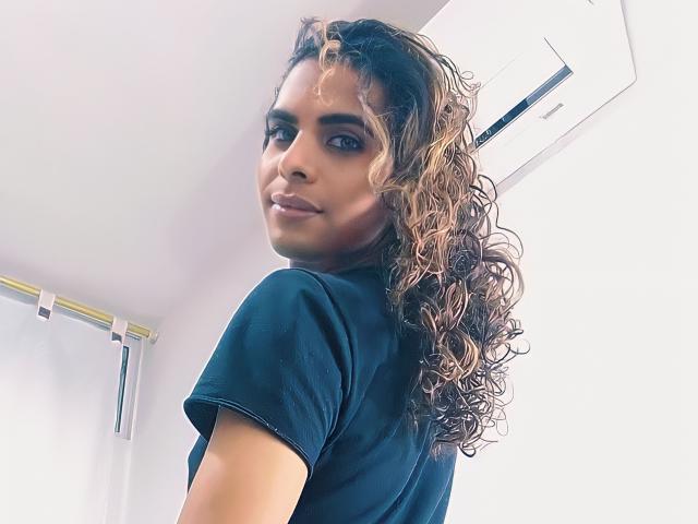 KarenSofia69, avec des cheveux bruns bouclés, un t-shirt noir, regardant en arrière, souriante, peau de couleur moyenne, yeux marrons, maquillage discret, mur blanc, unité de climatisation en arrière-plan..