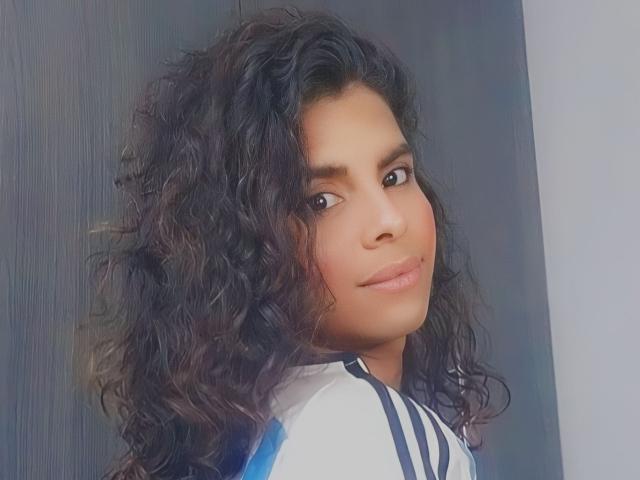 KarenSofia69, avec des cheveux noirs bouclés, des yeux marrons et une peau brun clair, portant une chemise rayée blanche et bleue, souriant doucement à la caméra..