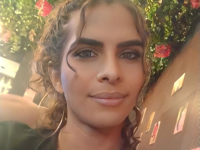 O rosto de KarenSofia69, com cabelo castanho ondulado, maquiagem discreta, brincos de argola, nariz pequeno, lábios rosados e cheios, um leve sorriso, luz do sol, folhagem verde com rosas cor-de-rosa ao fundo..