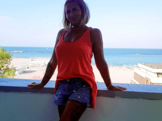 ChaudeEvely with tanned skin, medium boobs, wearing a red tank top and blue shorts with white studs, leaning on a blue railing, smiling, ocean and beach in background.