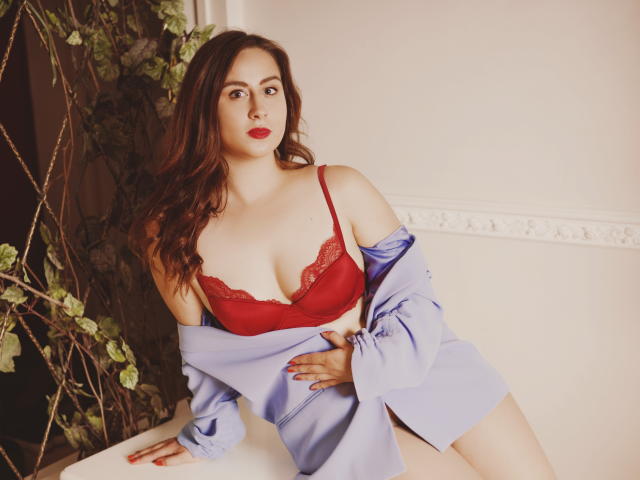 ViaLattea with red lace bra, showing her tits, has a blue shirt off her shoulders, long brown hair, and red lipstick, leaning on a white surface with green plants.