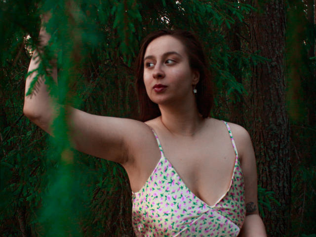 ViaLattea with medium tits, wearing a pink floral dress, standing in a forest, raising her arm, looking to the side, with greenery in the foreground.