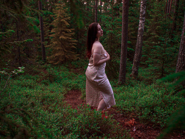 ViaLattea with long brown hair, wearing a white, off-shoulder dress, stands in a forest, holding her ass, looking back over her shoulder, surrounded by green foliage and trees.