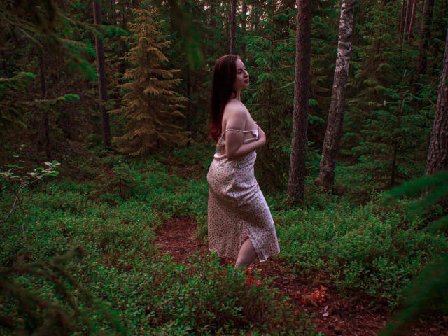 ViaLattea with medium tits, holding up a floral dress, exposing her ass, standing in a forest, looking back with dark brown hair, wearing glasses, in a green wooded area.