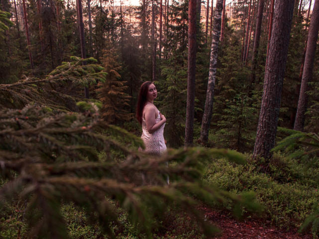 ViaLattea with long brown hair, standing in a forest, wearing a white lace dress, holding her tits, looking back, surrounded by pine trees and greenery at sunset.