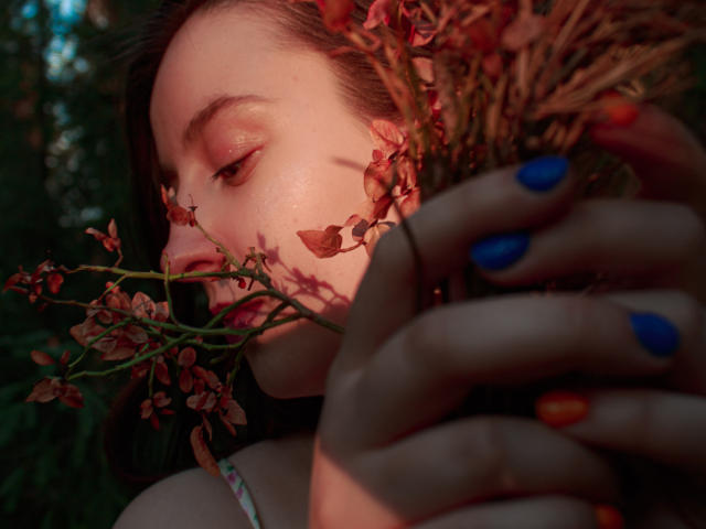 ViaLattea's face with pinkish-red petals in her mouth, blue, orange, and red painted nails, soft evening light, subtle pink eyeshadow, and brown hair, slightly blurred background.