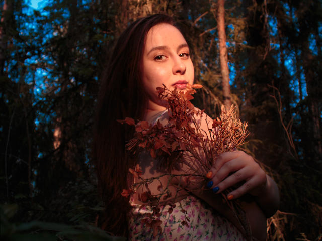 ViaLattea with brown hair, holding dried red leaves, sunlight on her face, blue nails, floral dress, forest background, looking slightly to the side, soft light, natural setting.