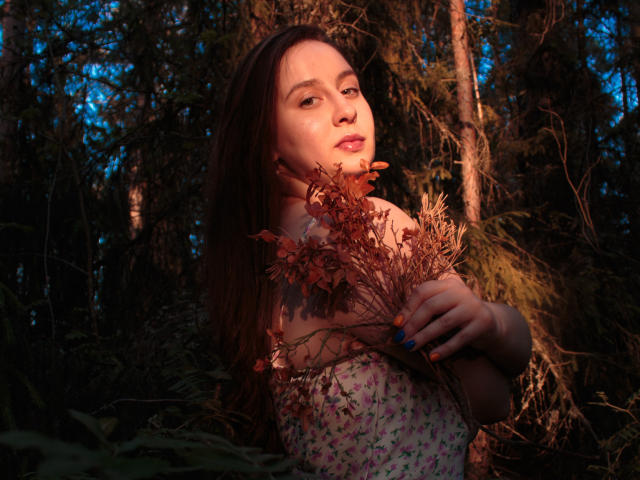ViaLattea with brown hair, holding dried flowers, wearing a floral dress, has orange nails, and a calm expression, in a dark forest with blue evening light.