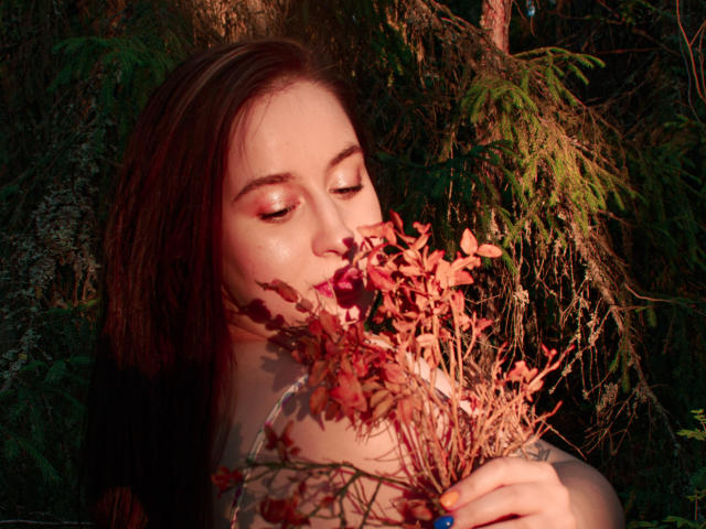 ViaLattea with red-brown hair, eyes closed, holding red flowers to her nose, sunlight on her fair skin, dark fur-like garment, forest background, soft golden hour light.