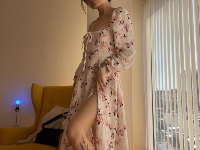 Tsukerberg in a floral dress with a thigh-high slit, showing a leg tattoo, standing by a window, holding her dress, with her hand on her pussy.