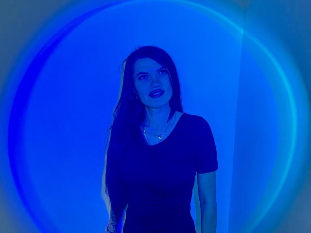 MilaLoona with blue-tinged skin, long dark hair, black shirt, standing, looking up with slightly parted lips, wearing a necklace, in a blue circular light effect.