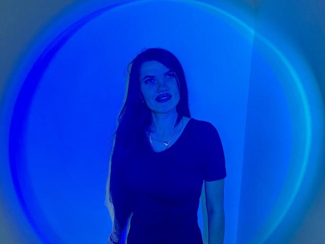 MilaLoona with blue-lit skin, dark wavy hair, black shirt, subtle smile, necklace, and upper body, standing in front of a circular blue light.