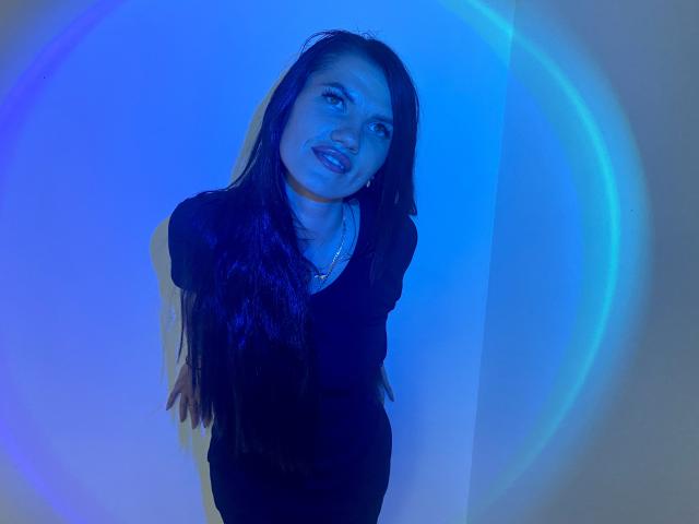 MilaLoona with long black hair, wearing a black dress, standing and leaning forward, blue light cast on her face, hands behind her back, slight smile, looking up.
