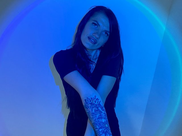 MilaLoona with long dark hair, black shirt, and tattooed arm, standing with arms crossed, blue lighting, looking at camera, slightly smiling, mouth slightly open.