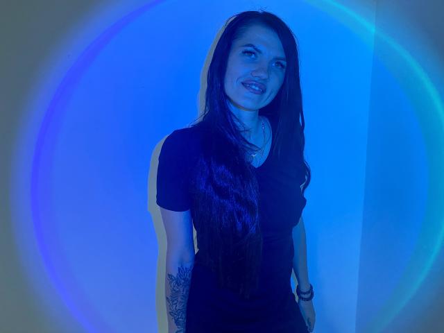 MilaLoona a de longs cheveux noirs, une chemise noire, des tatouages sur son bras gauche et un sourire. Elle se tient debout avec une lumière bleue circulaire derrière elle..