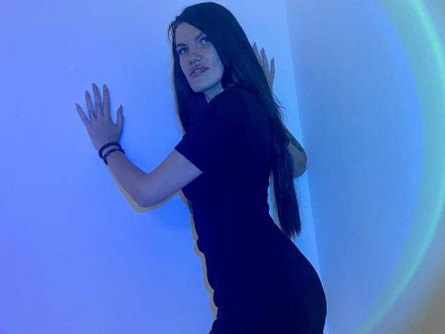 MilaLoona with long black hair, wearing a tight black dress, standing against a blue-lit wall, hands up, looking back with a slight smile, showing her ass.