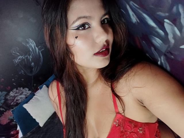 LucyDumont with long brown hair, red lipstick, and black heart makeup, wearing a red lace bra, showing her tits, looking at the camera, on a blue pillow.
