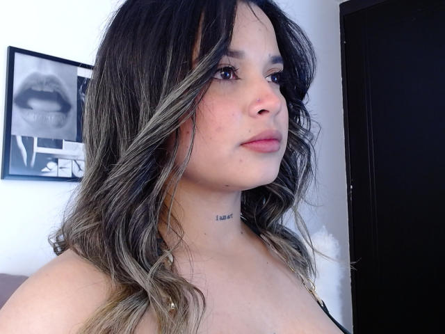KateZoe heeft een lichtbruine huid, golvend donkerblond tot bruin haar met een ombre-effect, en draagt geen top, met kleine borsten. Ze draagt een zwarte choker en kijkt naar rechts met een neutrale uitdrukking..
