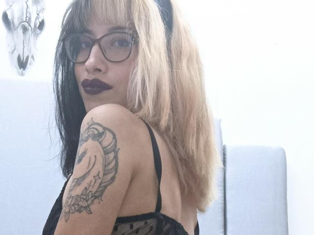 Laila Stewart, con cabello negro y rubio, gafas, lápiz labial oscuro, una blusa de encaje negro, mostrando un tatuaje en el brazo superior, mirando hacia atrás. Sus senos son parcialmente visibles..