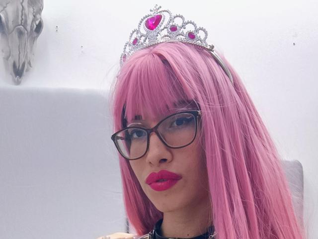 LailaStewart with pink hair, black glasses, and a silver tiara with a pink heart, has red lipstick and is looking at the camera. Boobs are not visible.
