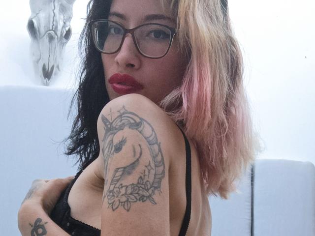 Laila Stewart, met een bril, rode lipstick, zwart-roze ombre haar, een zwarte bh, een tatoeage op de rechterarm, die haar linkerborst vasthoudt, met een schedel op de achtergrond, en die in de camera kijkt..