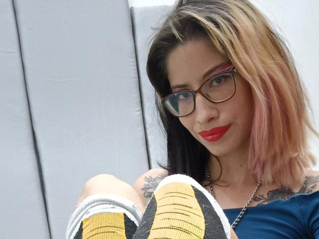 Laila Stewart, met roze en blonde haren, een bril, rode lippenstift, tatoeages, een blauw topje, die gele sneakers vasthoudt, zittend met de benen omhoog en die naar de camera kijkt..