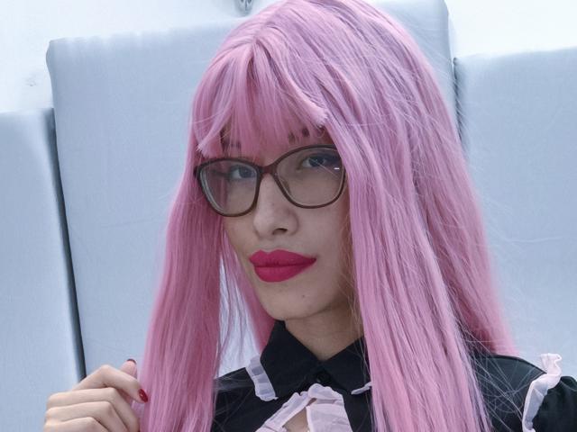 Laila Stewart con peluca rosa, gafas, lápiz labial rojo, atuendo blanco y negro, sujetándose el cabello, sonriendo, mostrando senos pequeños, piel clara, esmalte de uñas visible..
