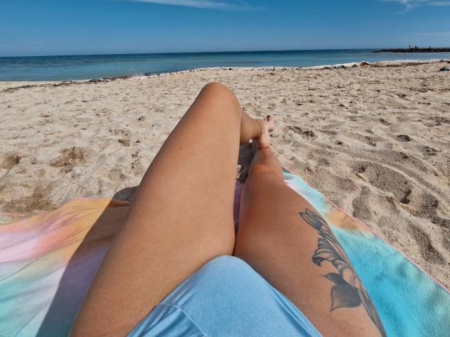 ValeriMair's tan thighs, with a colorful towel and a tattoo on the right thigh, lying on a sandy beach, legs crossed, feet touching, blue ocean and clear sky.