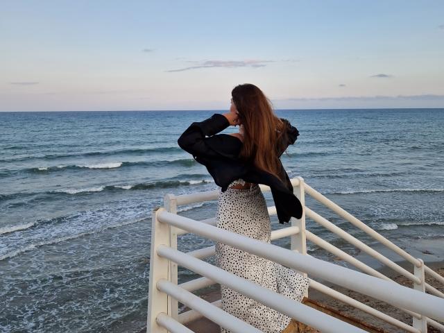 ValeriMair with long brown hair, black jacket, white polka-dot dress, standing on white staircase, holding black bag, facing ocean, small waves, clear blue sky, soft sunset light.